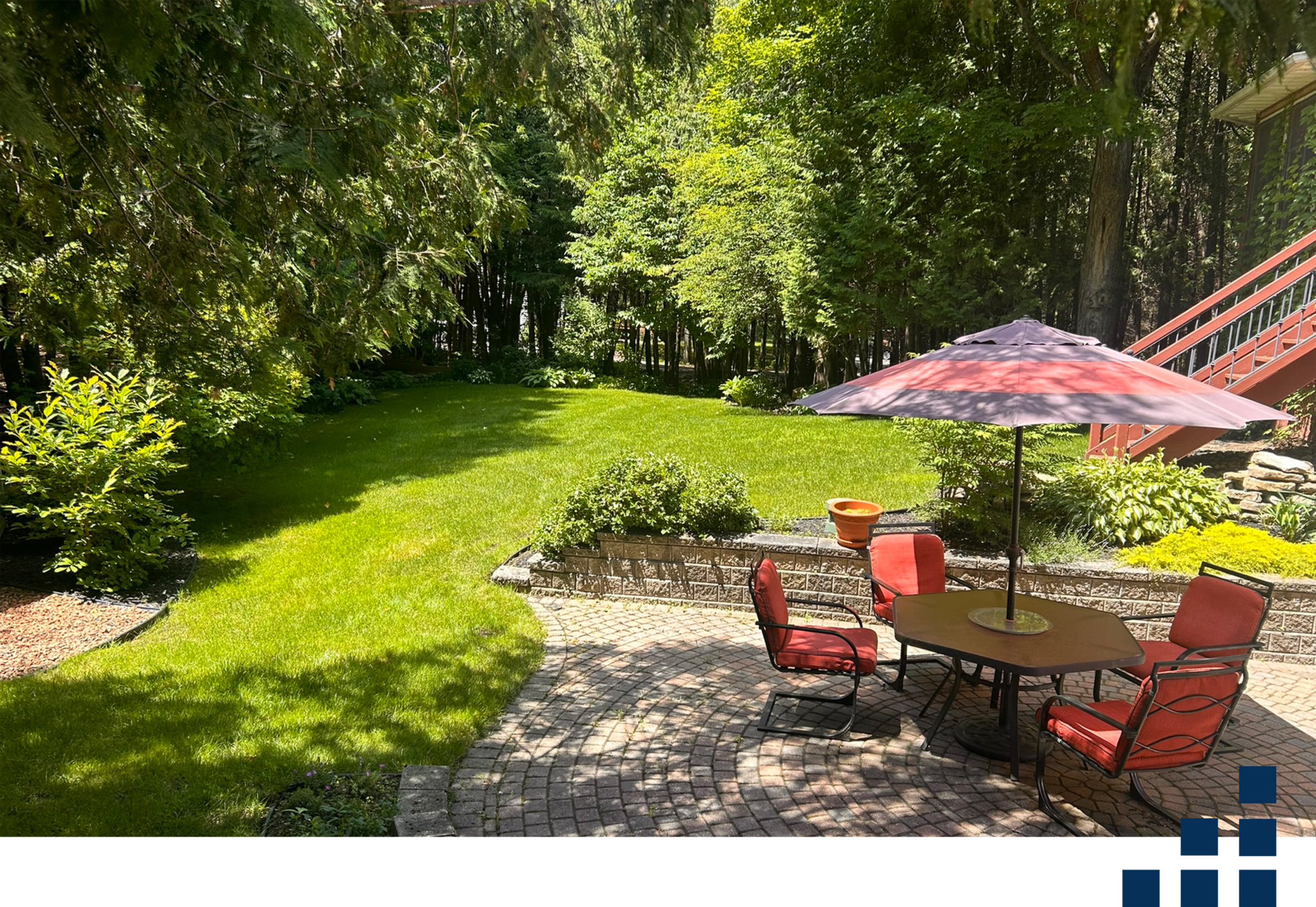a backyard with lush green grass, rimmed by trees for privacy. There is a brick landscaped patio with a retaining wall and a small patio set.